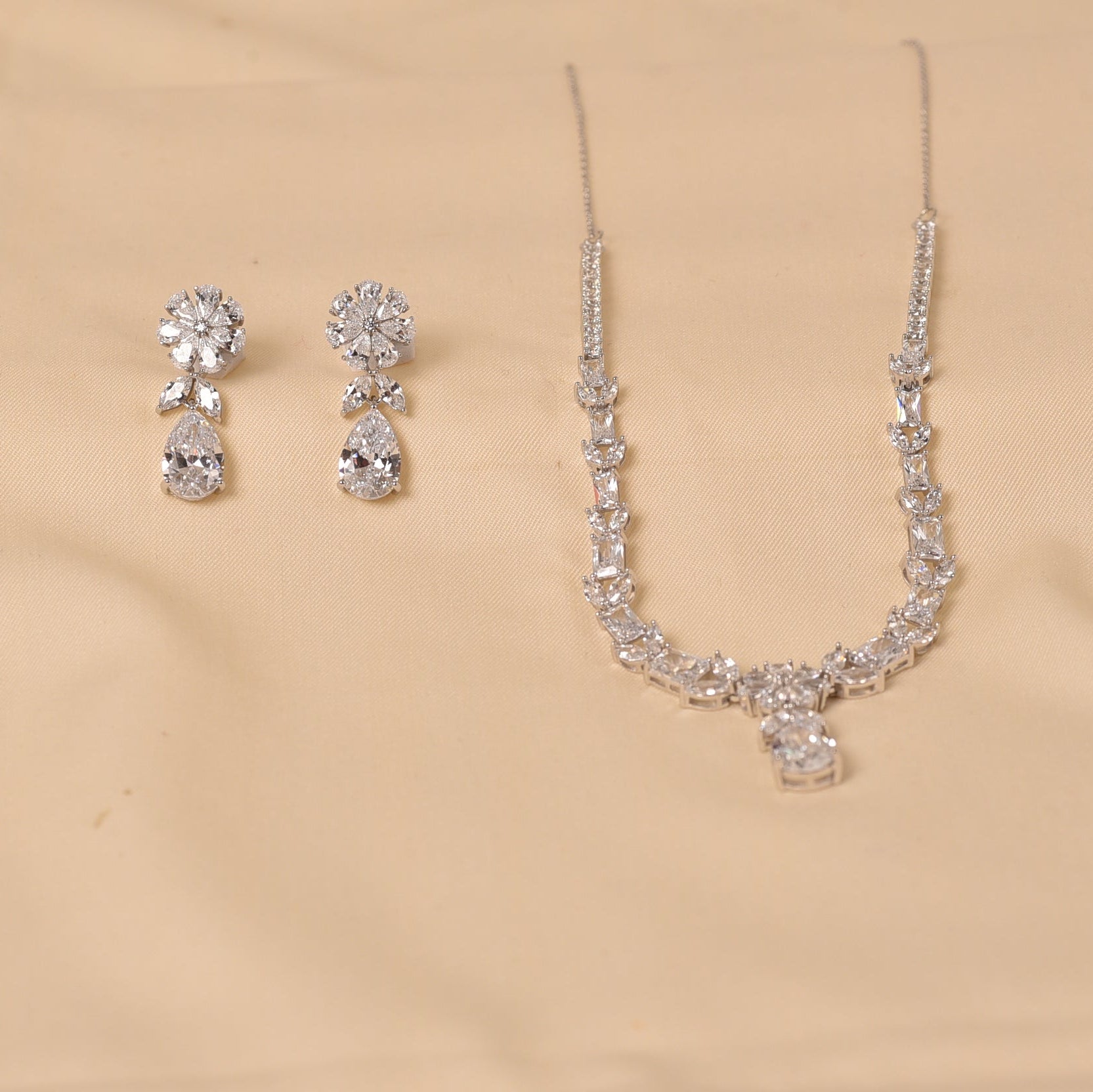 A silver-tone jewelry set featuring a delicate necklace and matching drop earrings, both adorned with clear crystals, displayed on a beige satin fabric background.