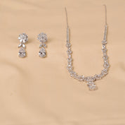 A silver-tone jewelry set featuring a delicate necklace and matching drop earrings, both adorned with clear crystals, displayed on a beige satin fabric background.