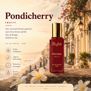 A 20ml bottle of Elyta Pondicherry Fruity Eau de Parfum displayed on a stone ledge in a scenic, sunlit street setting. The bottle is deep red with a shiny gold cap, and the background features warm-toned colonial buildings, hanging pink flowers, and white plumeria flowers.